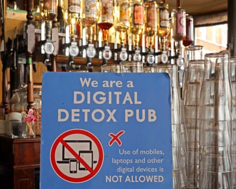 A blue sign in foreground on a pub serving counter reads: 'We are a digital detox pub' with bottles behind and stacked glasses to the right of photo