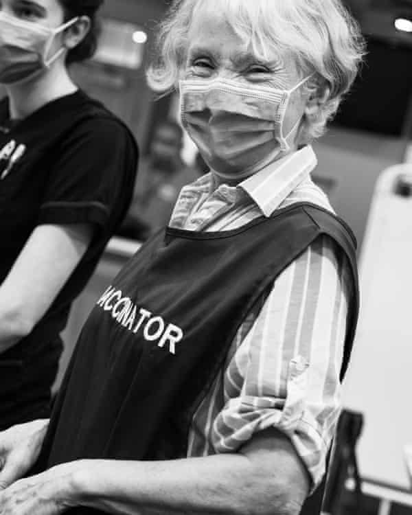 Black and white photo of a woman wearing an apron saying 'Vaccinator', looking at the camera and smiling behind her face mask, with another person in the background