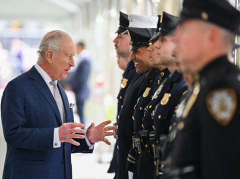 The king, in a blue jacket, speaks to a man in dress black, who is grinning, in a line of other people in dress black and caps, who are staring ahead.