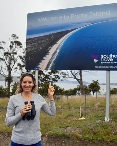 Dr Rochelle Steven is holding a pair of binoculars and pointing at a sign behind her which says ‘welcome to Bruny Island’