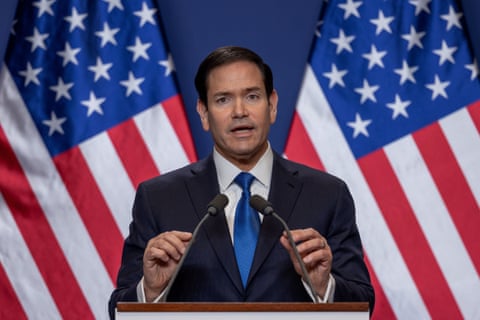 man in suit speaks into microphones, american flags behind him