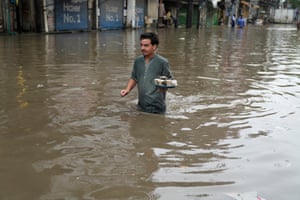 A man wades through waist-deep brown water carrying a small tray of coffee cups