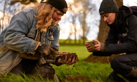 Ellie Bruce e Lucy Gray accovacciate su entrambi i lati di una buca scavata in un campo, tenendo in mano e fotografando la loro scoperta