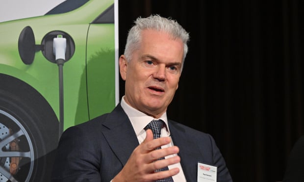 Paul Sansom is speaking and gesturing with one hand, in front of an image of a green electric car charging