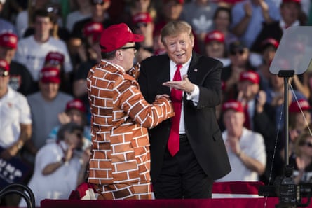 A man in a brick-patterned suit and Maga hat on stange with Donald Trump at a campaign rally.