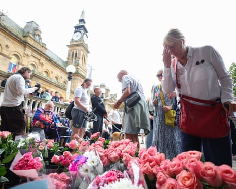 People gather around pink roses and flowers laid on the ground in Southport centre