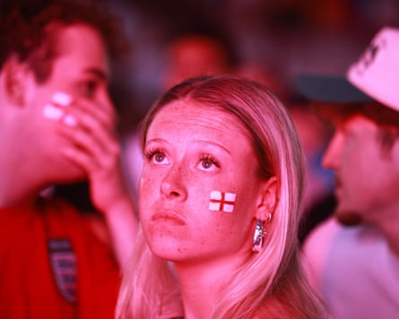 A young woman with England flags painted on her face looks sadly at a big screen.