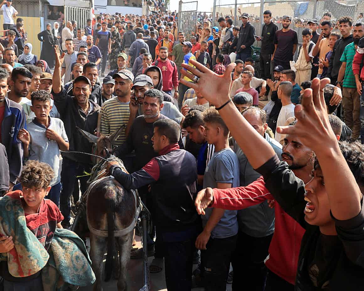 A crowd, mostly men and boys, gathers in the street; some look angry and have their arms raised; one young boy has a blanket over his shoulders and a man is leading a donkey through the people