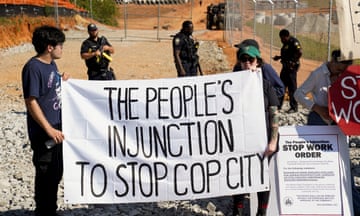 Two people hold a white sign reading 'The people's injunction to stop Cop City' as police officers stand behind them.