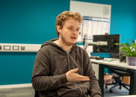 George Bate, 21, has short curly fair hair and a short light brown beard. He wears a grey hoodie and is sitting in a classroom-like room with a blue wall behind him. He holds up one hand as he speaks.
