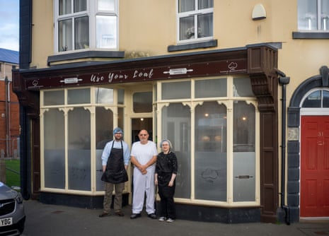 A young man and a young woman in aprons and hairnets stand outside an old-fashioned shopfront either side of an older man in a white baker’s outfit