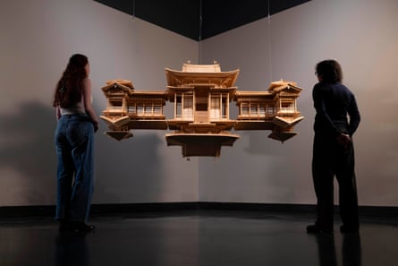 Two people stand in front of a wooden model of a shrine that is suspended from the ceiling