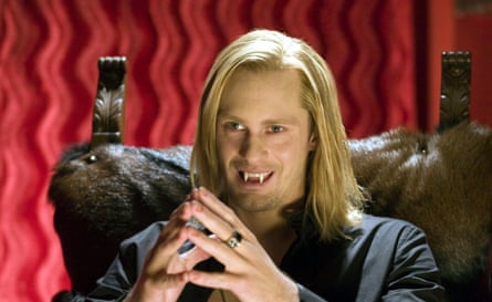 Head and shoulders of a young man with longish blond hair side parted sitting in an ornate leather chair with hands together as in prayer smiling and revealing fangs