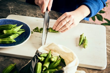 Hands holding a kitchen knife, cutting off the tops of okra pods on a white cutting board.