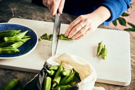 Tangan memegang pisau dapur, memangkas bagian atas buah okra di atas talenan putih.