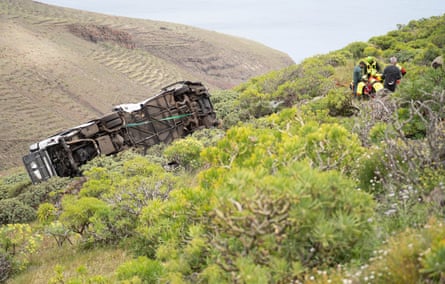 Bus on its side on a grassy slope with rescue workers nearby.