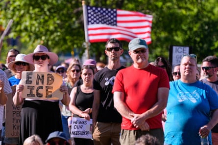 people with solemn faces stand outside at vigil, one person holding an american flag and others holding signs such as âÂÂend ICEâ and âÂÂhe was our neighborâÂÂ