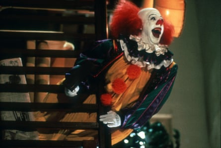 Pennywise the clown leans around a wooden staircase, mouth open wide, in IT