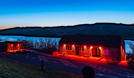 A low stone building glows with red lighting at dusk, overlooking a reservoir and hills