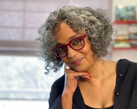 Arundhati Roy, with curly above-shoulder-level hair and wearing glasses and a button shirt over a T-shirt, rests her chin on her hand and smiles slightly