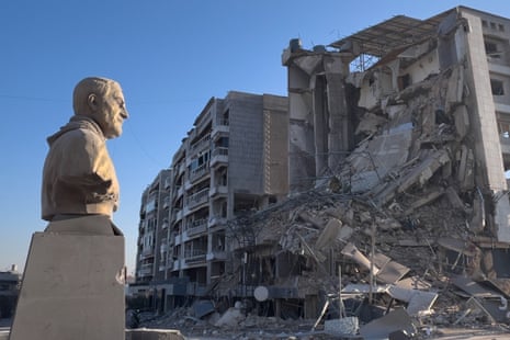 A stone bust of the late Iranian General Qassem Suleimani stands before the wreckage of a dsetroyed building, set against a blue sky.