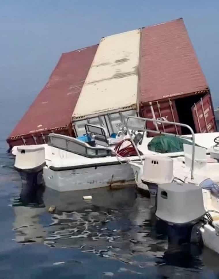 Small motorboats are pulled up alongside a large red and white shipping container which is part-submerged but mostly floating.