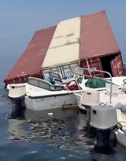 Small motorboats are pulled up alongside a large red and white shipping container which is part-submerged but mostly floating.