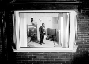 black and white photo of a man in a suit in a living room, photographed through a window from outside the house