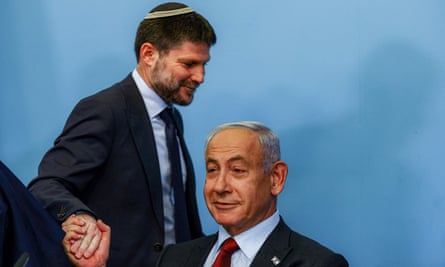 Smotrich clasps Netanyahu’s hand as he walks behind the prime minister, who is seated. Both are smiling