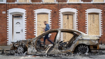 Burnt-out shell of a saloon car parked next to boarded-up homes