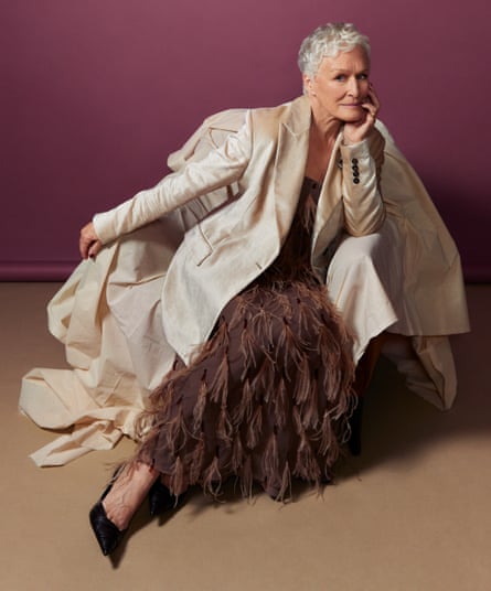 Glenn Close sitting elegantly on an armchair covered with cream material with her left arm leaning on the arm of the chair and her chin resting in her left hand, and her right arm stretched out across the other side of the armchair, wearing a smart cream blazer and full-length brown feathered skirt