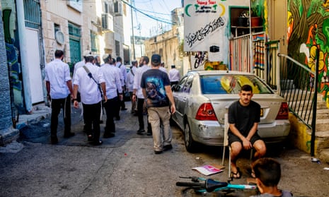 People walk down a street as a man sits on a chair at the side of the street