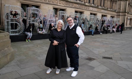 Shanaz Gulzar and Dan Bates stand in the city central square in front of letters spelling Bradford in fairy lights; volunteers are sitting in and standing by the letters. 