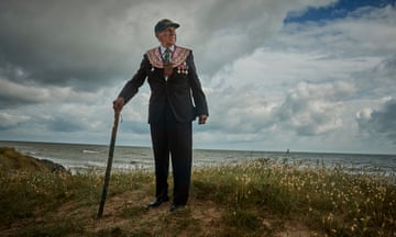 Man stands wearing his medals and leaning on a stick with sand and beach in background