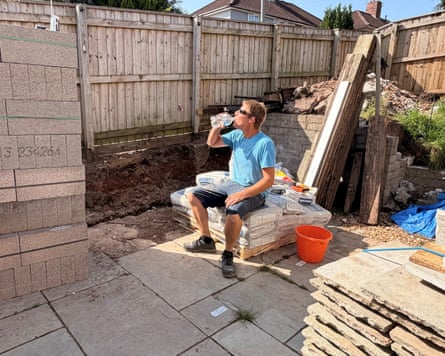 A man sits on paving slabs in a sunny garden drinking a bottle of water