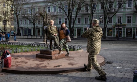 Two women pose on a sculpture of an old-fashioned chair in a public square, while a man in military fatigues takes a photo fo them