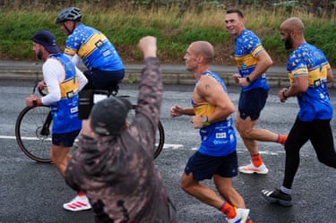 Jones-Buchanan and Sinfield run alongside two other men and a man on a bicycle as a person cheers them on from the side of the road