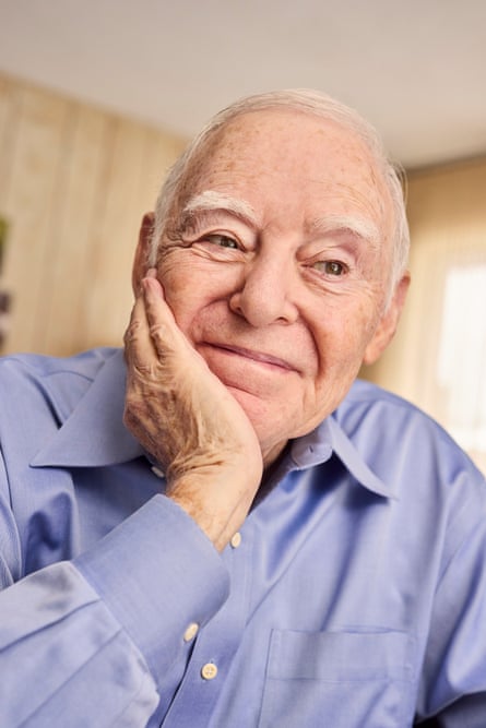 A man with short, white hair smiling while looking slightly off camera, with his head leaning on his right hand, wearing a blue shirt