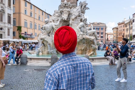 A man in a bright red turban and blue and white checked shirt stands in front of the fountain, a tall classical-style sculpture with nude figures, as others look on and take pictures