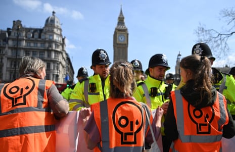 Three Just Stop Oil protesters wearing orange hi-vis jackets speak with police in front of the Houses of Parliament