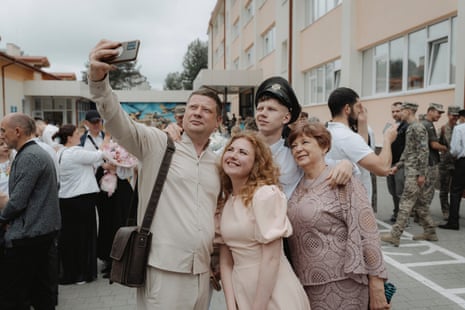 a young man in a black cap and white shirt stands with his arm around an older lady in a long-sleeved light brown lace top and skirt and behind a woman with reddish-blond curly hair who may be his mother, who wears a peach-coloured dress with puffy short sleeves, while a man who may be his father, wearing a pale cream shirt and trousers, holds up a camera.