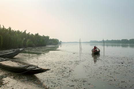 A man sets out in a small flat-bottomed boat on a wide and placid river with dense palms and other vegetation on the banks