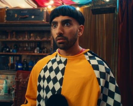 A still from the BBC surreal comedy series Juice, featuring the creator and star Mawaan Rizwan as the character Jamma. He is a young man with a distinctive black bowl-cut hairstyle and a short beard.