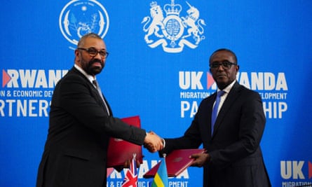 UK home secretary James Cleverly and Rwanda foreign minister Vincent Biruta, each holding a folder, shake hands in front of small flags from each country and a poster reading “UK Rwanda migration & economic development partnership”