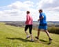 Two people jog along a grassy hilltop path with rolling countryside visible below