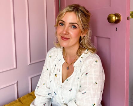 she is in her early 30s with long blond hair tied back from her face, and blue eyes; she wears a white cotton blouse or dress and is sitting on a mustard-coloured sofa or cushion in front of pale pink wooden panelling and a brighter pink wooden door with brass doorknob.