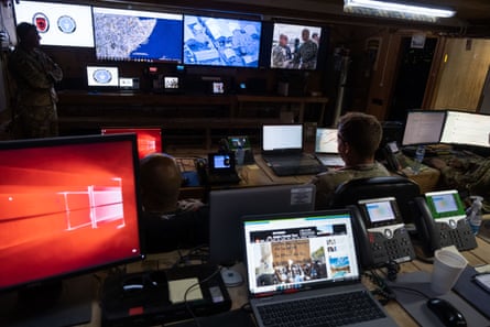 Banks of screens and computers connected desks and nan wall, pinch uniformed men watching them.