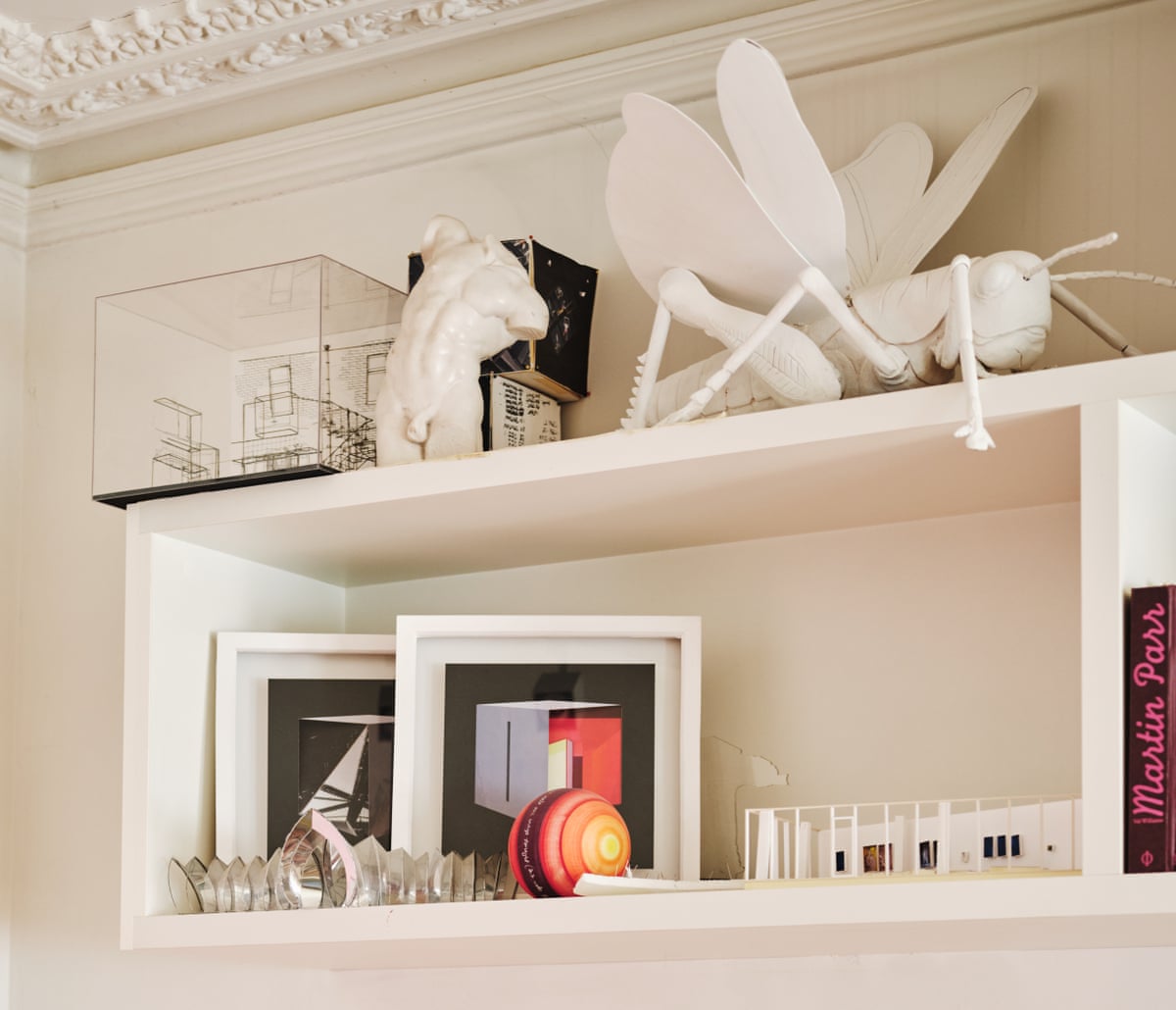 A white shelf on a white wall containing models and framed pictures