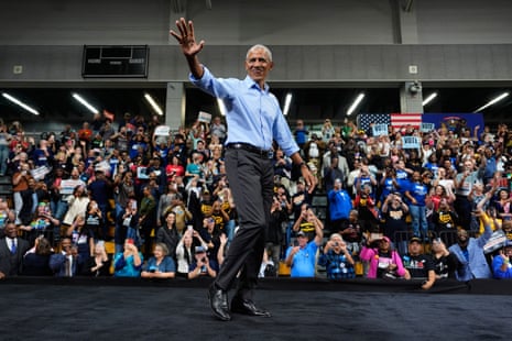 Barack Obama, in blue button-up shirt, walks on platform waving to animated crowd.