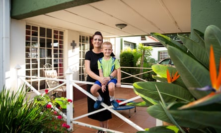 A little boy wearing a green t-shirt and blue shorts sits on a fence, held by a smiling woman